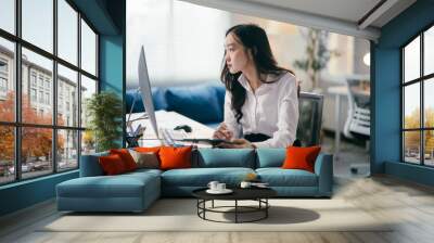 Focused young woman working at tidy office desk, reviewing laptop info and jotting notes, surrounded by tech and paperwork. Successful business career vibes Wall mural