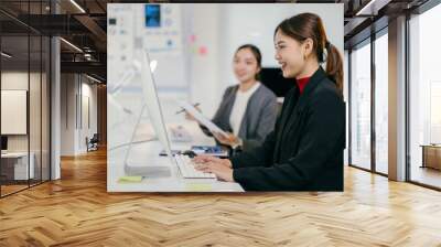 Two asian businesswomen are working together in a modern office setting, collaborating on a project. One businesswoman is typing on a desktop computer while the other reviews documents Wall mural