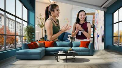 Two joyful asian coworkers clapping in celebration of successful brainstorming in their office, beaming with happiness and teamwork. Modern workspace filled with creative ideas Wall mural