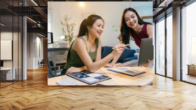 Two young asian businesswomen collaborating in a modern office, working on a startup project with laptops and documents. Happy, confident, and professional, they discuss strategies for success Wall mural