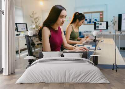 Two young businesswomen are concentrating on working at their laptops in a bright modern office, surrounded by paperwork and digital devices Wall mural