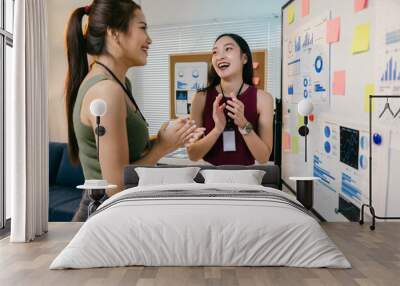 Two young businesswomen are standing in an office with a whiteboard covered in graphs and charts, clapping their hands and smiling Wall mural