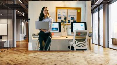 Young professional woman holding a laptop while leaning on a desk in a modern office, surrounded by computers and whiteboards with data visualizations Wall mural