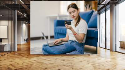 Young woman is sitting on the floor using a laptop and holding a credit card, making an online purchase from her living room Wall mural