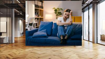 Young woman sits alone on her sofa at home, feeling sad and overwhelmed with unhappiness, stress, and worry, reflecting on her problems and facing feelings of loneliness and anxiety Wall mural