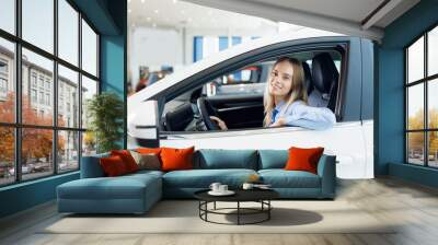 Portrait happy young woman driver behind wheel of new car on test drive before buy in showroom Wall mural