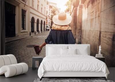 Young woman walks with bag and in straw hat tourist street sun rays along outdoors of old town Wall mural