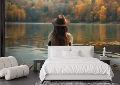 Back view of fashioned young woman sitting on wooden dock looking at lake view. Female hipster with brown hat relaxes on the edge of jetty admiring autumn colors. Wonderful nature getaway Wall mural