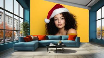 Young woman with curly hair wearing a Santa hat posing against a yellow background. Wall mural