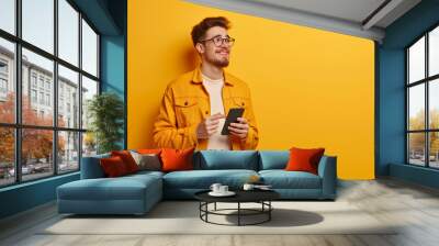 Portrait of handsome happy young man holding smartphone isolated on yellow background. Studio shot Wall mural