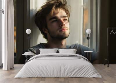 Young man with a contemplative expression sitting by a window with natural light Wall mural