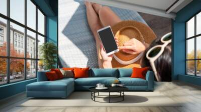 Asian woman sitting on sunbed near swimming pool using mobile phone. Young traveler female working on cellphone during her summer vacation trip. Technology and lifestyle concept. Copy space, top view Wall mural