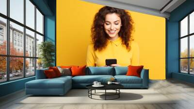 Beautiful young brunette woman holding a phone on yellow background Wall mural