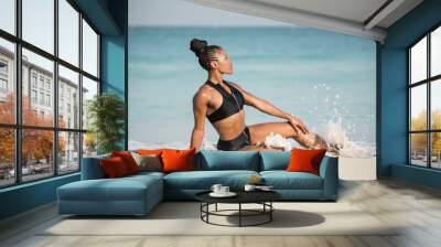 Beautiful African American Black female fitness model on the beach wearing black sportswear sitting on the waters edge being splashed by a small wave.
 Wall mural