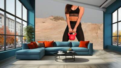 Beautiful brunette fitness model wearing black tights and black sports bra  exercising in a hot desert area on a bright sunny day with red kettle bell Wall mural