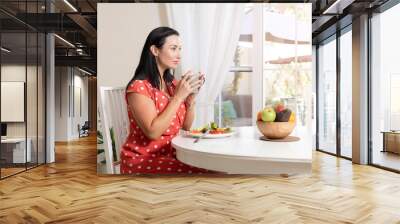 Beautiful brunette woman sitting wearing a red and white polka dot dress at dining table eating a healthy salad and drinking coffee or tea with a bowl of fruits on table with garden in the background Wall mural