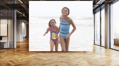 Two mixed-race sisters playing and laughing having fun on the beach on a bright tropical summer holiday Wall mural