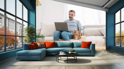 Portrait of senior old caucasian man stay home sit on the sofa  living room working on computer with lovely dog. Happy moment of pure love middle age male playing with little dog in winter Wall mural