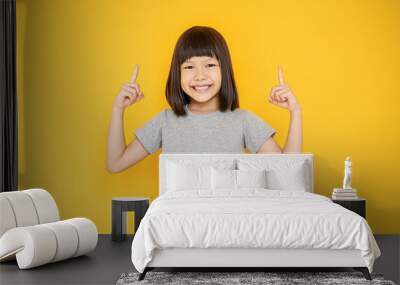 Portrait of young fun smart happy little cute asian girl isolated on yellow with copy space studio shot. Education for elementary kindergarten, little girl finger point up back to school concept Wall mural