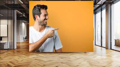 Smiling young man in white t-shirt looks to the side and point finger. Cheerful man isolated on orange background, studio shot for product advertisement. Wall mural