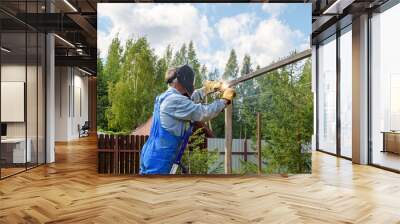 Man welder in a welding mask, construction uniform and protective gloves cooks metal on a street construction site. Construction of a pavilion, pergola near a country house on a summer day. Wall mural