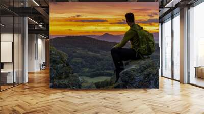 Thinking man sit on rock. Man on exposed summit watching to misty far distance. Cold day in mountains. Red Sunset setting behind clouds Wall mural