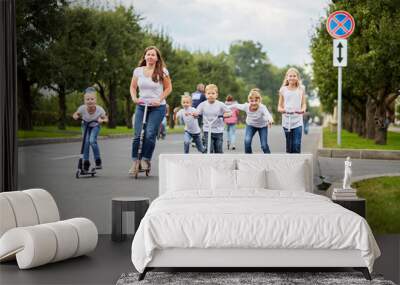 Woman and five children on scooters, roller skates and skateboard ride on street Wall mural