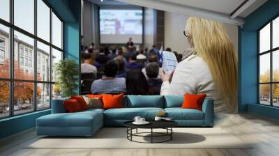 Young blond woman stands looking into booklet in auditorium full of people, shallow dof. Wall mural