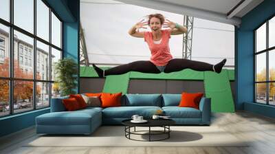 Young woman jumps on trampoline attraction making leg-split and grimacing by face Wall mural