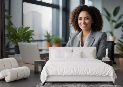 A businesswoman in a grey suit sits at her desk in a modern office, working on her laptop with a confident smile Wall mural