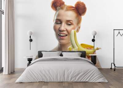 Red-haired girl with a banana in her hands. Holds a banana next to his face and lips. Isolated on a white background. Wall mural