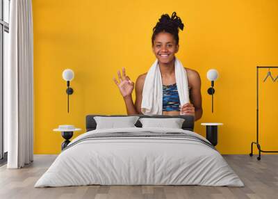 Young happy dark skinned teenager girl, athletic body isolated on a yellow background. African woman smiles, in a sports uniform holds towel on neck, shows ok sign after fitness. Copy space. Wall mural