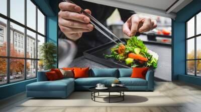 A close-up of hands wearing black gloves carefully plating a gourmet salad with mixed greens, sprouts, and colorful vegetable slices on a dark background Wall mural