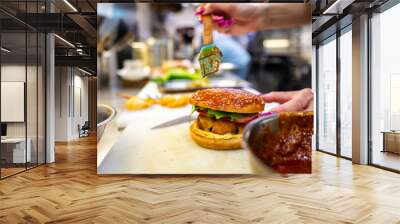 Close-up of a hand applying sauce to a burger with a brush in a kitchen setting. Ingredients and fries are visible in the background, emphasizing the culinary preparation process Wall mural