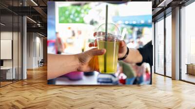 Close-up of two hands exchanging a plastic cup with a yellow-green beverage and a straw against a blurred outdoor background Wall mural