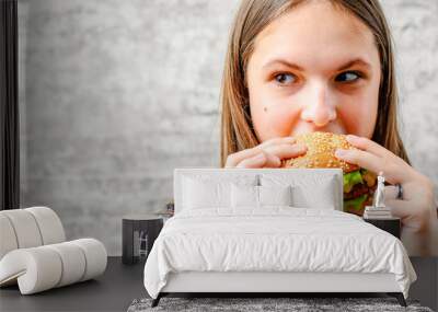 portrait of young teenager brunette girl with long hair eating burger. Girl trying to eat fast food on gray wall background Wall mural