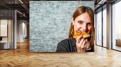 portrait of young teenager brunette girl with long hair eating slice of pizza on gray wall background Wall mural