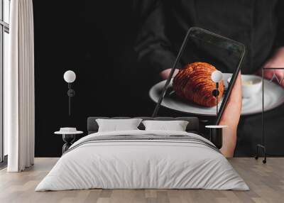 woman hand holding and showing smart phone takes a photo chef in a black suit holds in his hands plate with croissant on Dark grey black background. food photos Wall mural