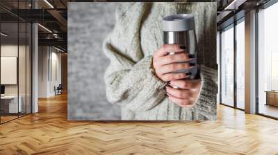 woman in a sweater holds a thermomug with a hot drink in her hands on gray wall background Wall mural