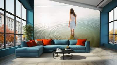 Woman in summer dress standing on seashore and looking at horizon. Young beautiful girl standing in water Wall mural