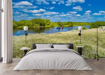 Men at kayak trip on blue river landscape near field of white daisy flowers, green forest with trees blue water clouds sky Wall mural