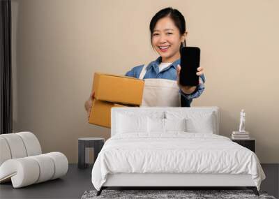 Entrepreneur asian woman wearing aprons on light brown background. She holding cardboard boxes and showing blank screen smartphone. Wall mural