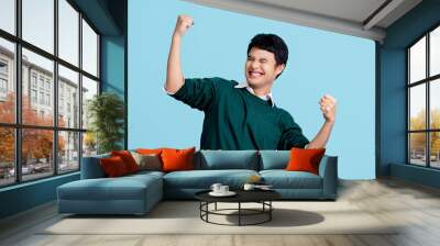 Portrait of a cheerful young Asian man in casual clothes. He raised his fist with a happy smile on his face as a gesture of celebration. Wall mural