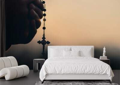 Man's hands pray to hold a bead rosary with Jesus Christ on the cross or the Crucifix with sunset. Christian Catholic religious faith holding a black rosary and praying. Wall mural