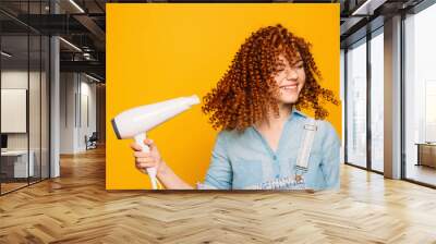 curly red-haired woman using hair dryer on yellow background. Making perfect curls Wall mural