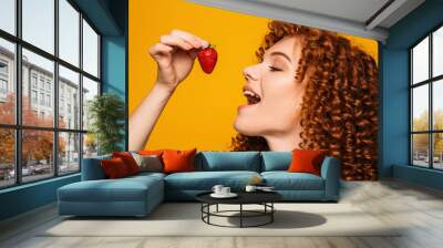 curly red-haired woman with open mouth wants to eat delicious strawberries on a yellow background. Wall mural