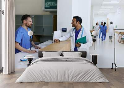 Indian doctor talking to male nurse near reception desk at modern hospital while working day. View of lobby medical clinic with patients and medical personnel Wall mural