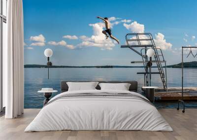 Side view of a teenage male jump diving from a diving tower with blue sky and horizon in the background. Wall mural