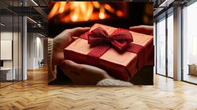 Close-up of two hands exchanging a beautifully wrapped red gift box with a shiny ribbon, set against a warm and cozy fireplace backdrop Wall mural