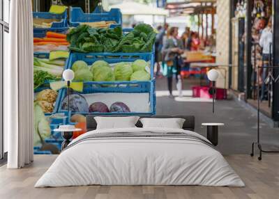 Outdoor selected focus view of various vegetables sell on row of blue plastic basket in front of stall outside grocery store beside corridor of open air market with blur background of people. Wall mural